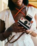 woman wearing white dress with small black polka dots holding white polaroid camera on a brown leather strap