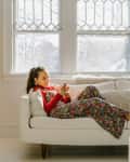 Woman in floral dress lounging on a white sofa, using a smartphone, with a yellow lamp and wooden dresser nearby.
