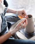 Person painting a clay pot they've made on a pottery wheel. Their hands are covered with drying clay.