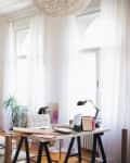Bright living room with white sofa, wooden desk, laptop, and large windows with sheer curtains.