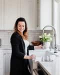Woman in a white kitchen smiling while washing dishes at the sink, with white cabinets and a vase of tulips nearby.