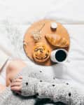 Person in cozy sweater reading a book on bed with coffee, croissant, cinnamon roll, and flowers on a wooden tray.