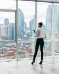 Person in business attire standing by large windows overlooking city skyline.