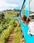 Woman leaning out of a blue train window, enjoying scenic green hills and wildflowers.