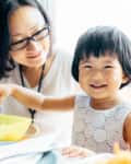 Woman and child smiling at a table, child holding a spoon over a yellow bowl.