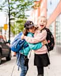 Two children with backpacks hugging and laughing on a city sidewalk near a glass building.