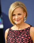 Woman in floral black and pink dress with short blonde hair and dangling earrings, smiling at an event.