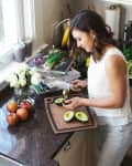 Woman slicing avocados in a kitchen with apples, flowers, and a spiralizer on the counter.