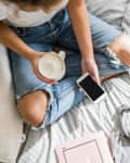 Person in ripped jeans holding a coffee mug and smartphone, sitting on a bed with a gray blanket and glasses nearby.