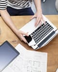 Person using a laptop and smartphone at a wooden table with a salad, tablet, and architectural plans.