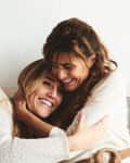 Two women smiling and embracing on a sofa, wearing cozy sweaters.