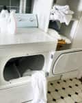Laundry room with washer, dryer, detergent bottles, and open cabinet with folded towels on a tiled floor.