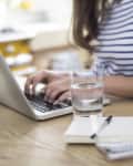 Person in striped shirt typing on a laptop at a wooden table with a glass of water, notebook, and blue floral vase.