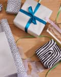 Gift wrapping supplies on a wooden surface, featuring black and white patterned paper, ribbons, and a blue bow.