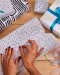 Hands wrapping gifts with black and white patterned paper, surrounded by ribbons and tape.