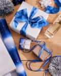 Gift wrapping scene with blue and gold paper, ribbon, and various wrapped presents on a wooden surface.