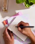 Person writing on a notepad surrounded by notebooks, a stapler, a plant, and a calculator on a desk.