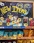 Display of new snack items in a grocery store, featuring colorful packaging, signage, and a shopper in the background.