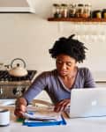 Woman working at a kitchen counter with a laptop, papers, and a mug, surrounded by kitchenware and decor.