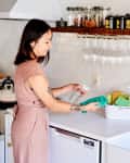 Woman cleaning kitchen counter with spray bottle and cloth, near gold faucet and shelf with jars and glasses.