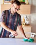 Man cleaning a white table with a spray bottle and cloth in a bright room with plants and shelves.