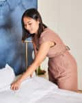 Woman in a brown dress making a bed with white sheets against a blue abstract wall.