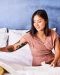 Woman in a pink jumpsuit arranging bedding on a white bed with a blue wall and a modern lamp in the background.