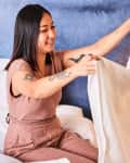 Woman folding a white towel on a bed with blue wall and bedside lamp.
