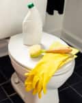 Toilet with cleaning supplies: yellow gloves, brush, and bottle on lid, black tiled floor.