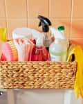 Wicker basket with cleaning supplies, spray bottles, sponges, and yellow gloves on a toilet tank against peach tiles.