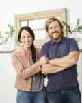 A smiling couple stands in a living room with bookshelves, plants, and a rustic mirror.
