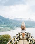 Person with a backpack standing on a wooden platform overlooking a lake and mountains.