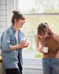 Two women laughing in a kitchen, holding mugs, near a window with a wooden countertop and white tiled walls.