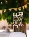 Cake topper reading "Hooray for the Happy Couple" with flowers and bunting in the background.