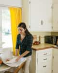 Woman unpacking dishes in a kitchen with yellow curtains, white cabinets, and a countertop with a toaster oven and kettle.