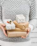 Person in a patterned sweater holding wrapped gifts in brown and white paper with twine.