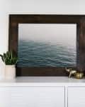 Framed ocean photo on white cabinet with potted plants, books, and brass elephant figurine.