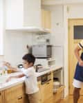 Man in apron setting table with fruit while child washes hands in kitchen.