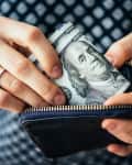 Hands holding a black wallet with rolled hundred-dollar bills.