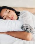 Woman sleeping peacefully on a white bed, wearing a white top with black embroidery.