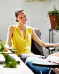 Two women having a conversation in a bright office with plants and stacks of papers on the desk.