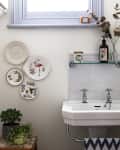 Bathroom with wall-mounted sink, decorative plates, plants, and a shelf with bottles and a candle.