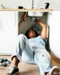 Person fixing a kitchen sink, lying on the floor with tools and pipes nearby, next to white cabinets.