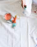 A painter kneeling on a white drop cloth with three open cans of paint and paintbrushes