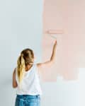 Person painting a wall light pink with a roller, wearing a white tank top and jeans.