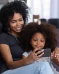 Happy smiling african american mother and little daughter taking selfie on smartphone, mixed race mom and child girl making video call on cellphone, mommy kid watch cartoons using phone app at home