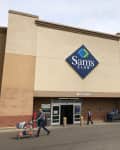 The exterior of a Sam’s Club store with a shopper walking in and away from it.