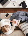 Woman sitting on sofa writing in notebook, with laptop, iPad, and phone nearby
