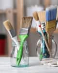 a variety of used paint brushes in glass jars. a pair of gloves sits on the table next to them