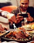 Front view of a young mixed race man sitting at a table for Thanksgiving dinner at home with friends carving the turkey, a young mixed race male friend taking a photo with his smartphone in the
